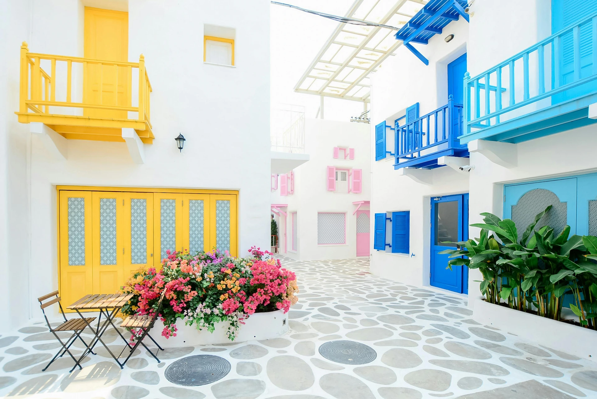 Bright alley with white buildings and colorful doors and balconies in yellow, blue, and pink, with a flower planter and a cafe table set.