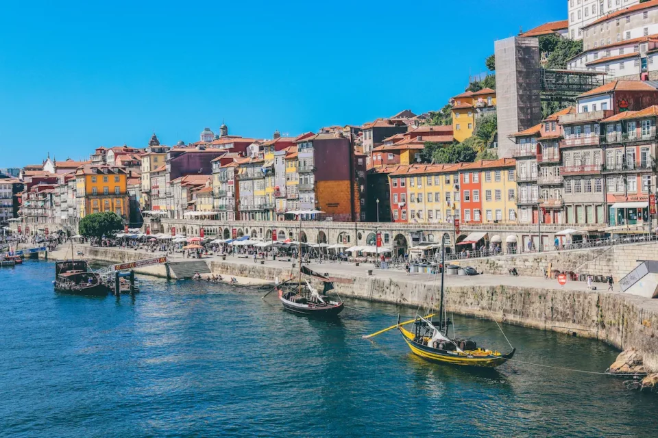 Colorful buildings along the Ribeira riverfront in Porto with traditional boats floating on the Douro River under a bright blue sky.