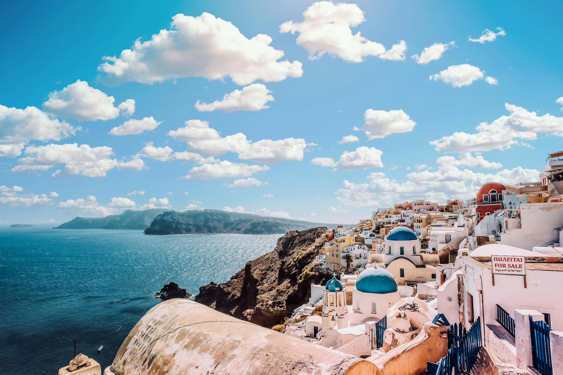 Scenic coastal village with white buildings and blue domes overlooking the Aegean Sea under a bright blue sky with clouds.